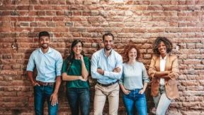 A group of five smiling people are leaning against a red brick wall.